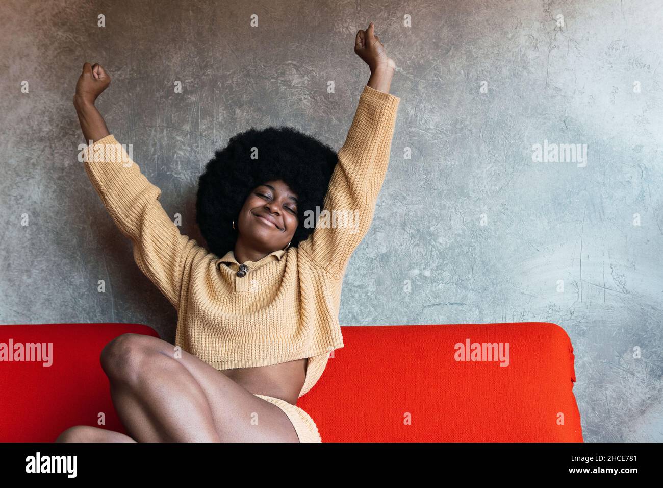 Entzückte afroamerikanische Frau mit Afro-Frisur und geschlossenen Augen, die sich mit erhobenen Armen in Heimkleidung streckte, nachdem sie zu Hause dagegen aufwachte Stockfoto