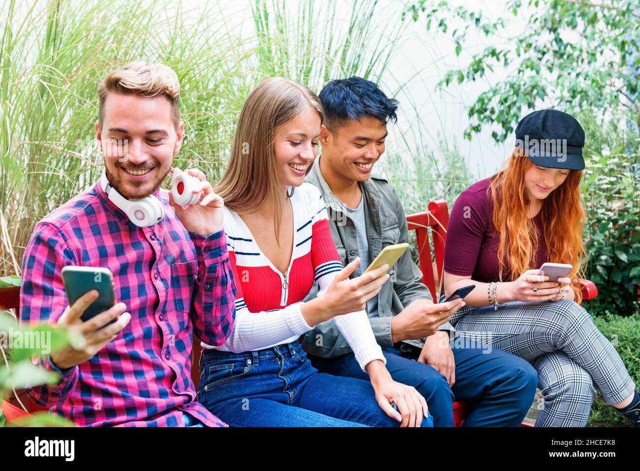 Fröhliche, multiethnische Freunde tragen stilvolle, legere Kleidung, sitzen auf der Bank und surfen auf der Bank im Park durch Mobiltelefone Stockfoto