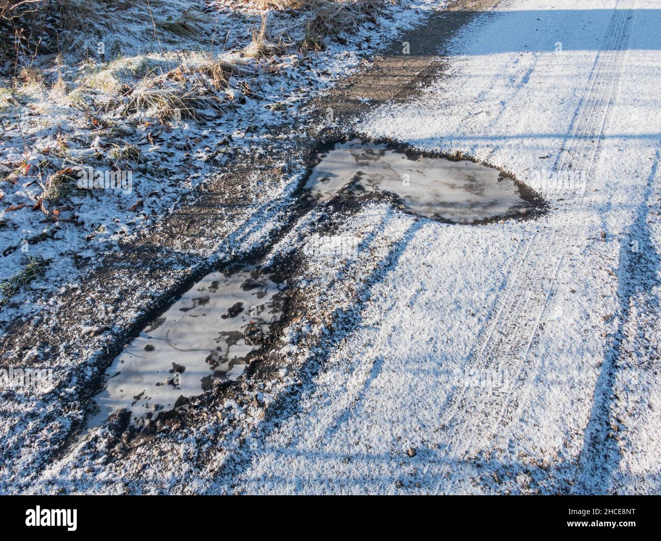 Unbefestigte, unbefestigte Straße. Es ist ein Wintermorgen. Der Boden ist mit einer Schneeschicht bedeckt, das Wasser in der Pfütze ist gefroren. Stockfoto