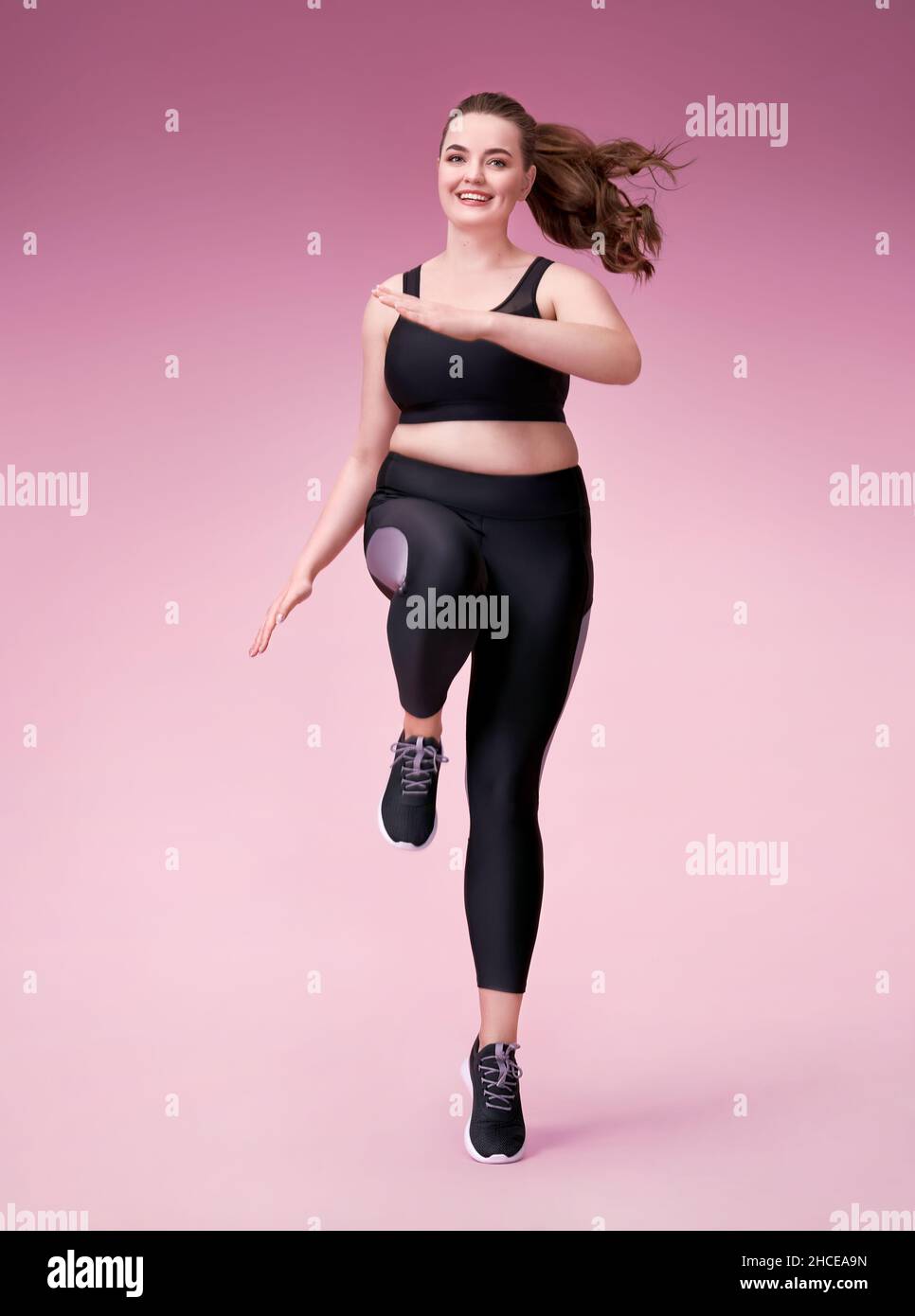 Sportliche Mädchen springen, tun Übung. Foto des Models mit kurviger Figur in modischer Sportbekleidung auf rosa Hintergrund. Dynamische Bewegung. Seitenansicht. Sport Stockfoto