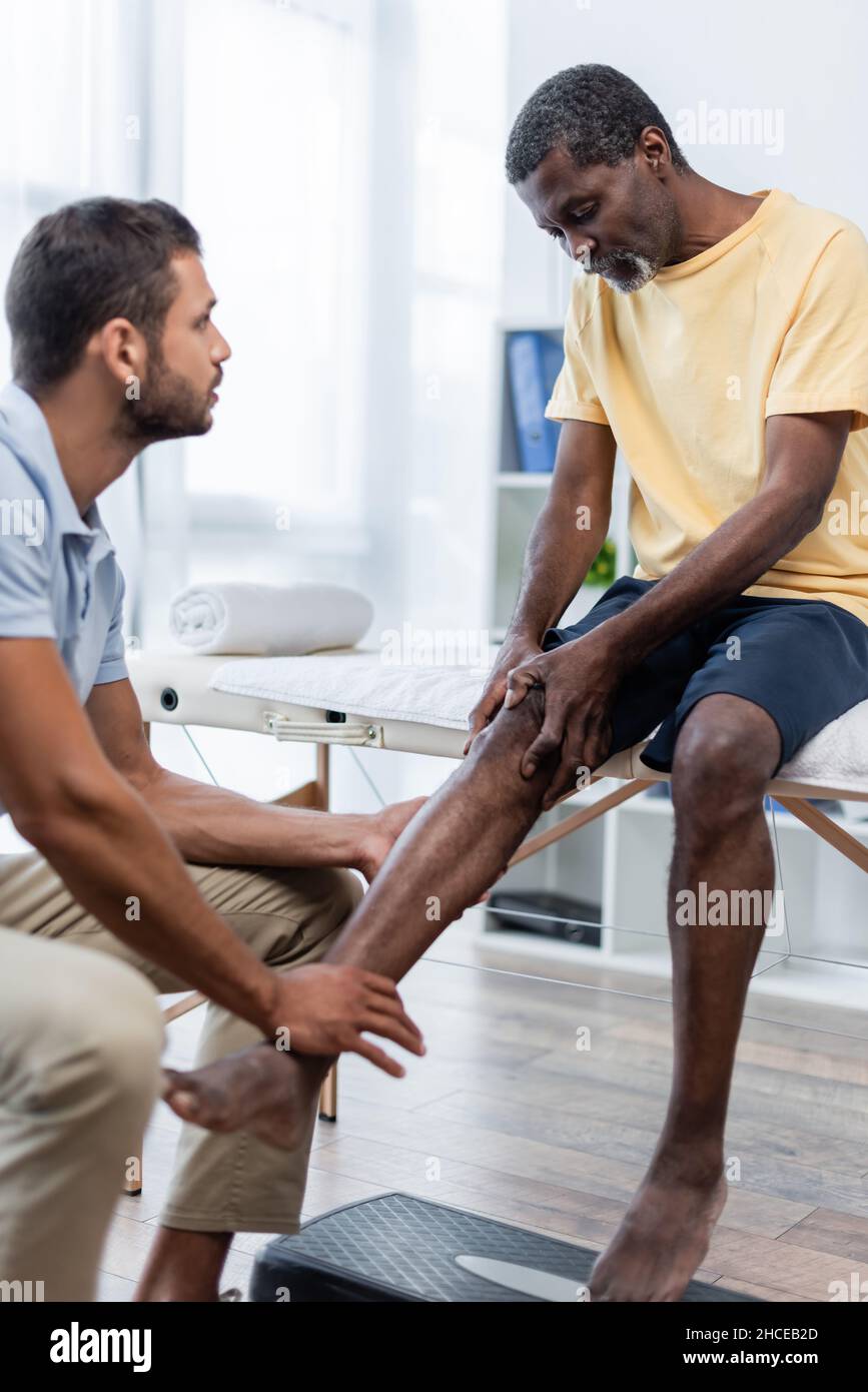 Reifer afroamerikanischer Mann berührt Knie, während er mit dem Arzt im Rehabilitationszentrum spricht Stockfoto