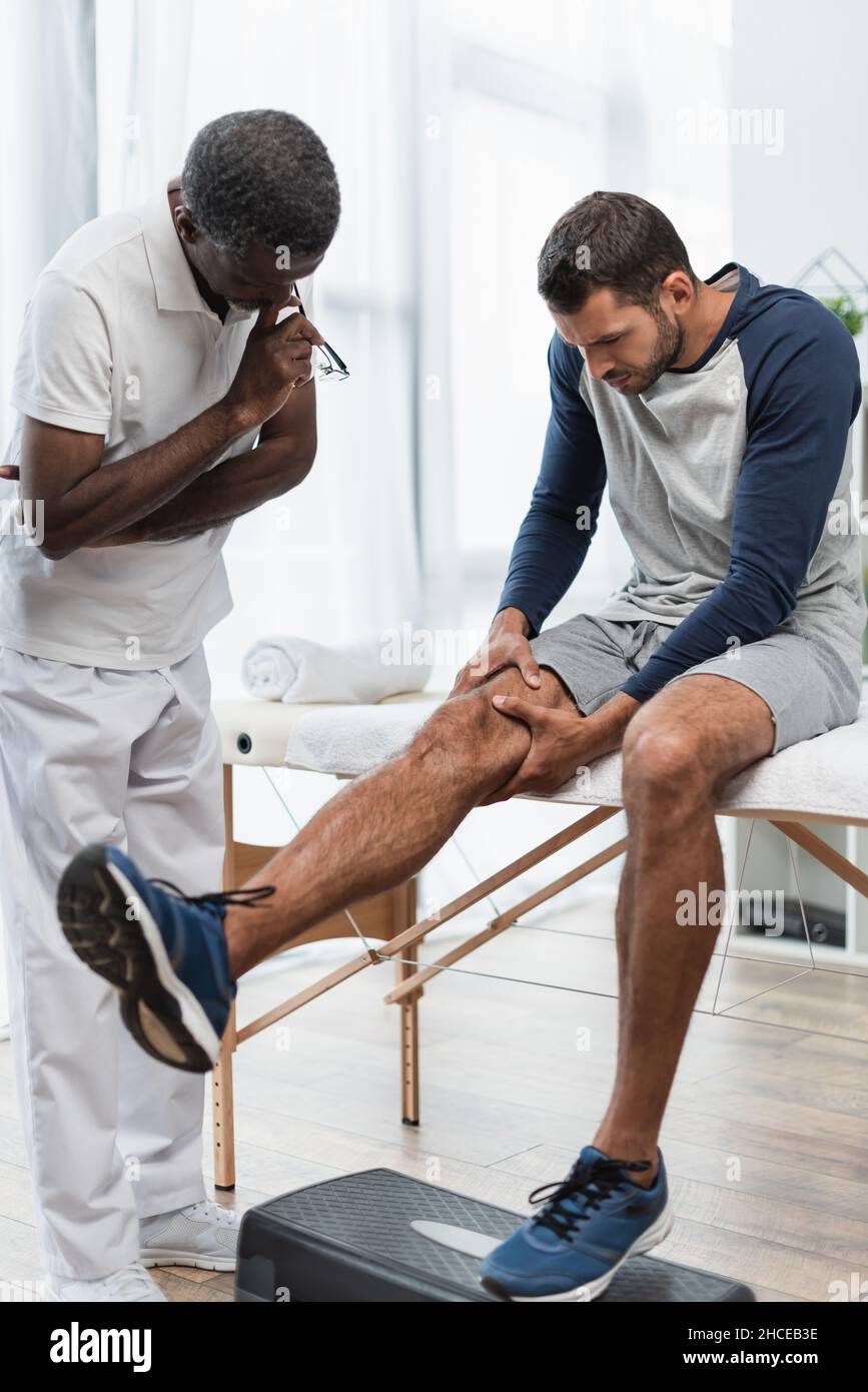 Der junge Mann berührt das Knie, während er auf dem Massageliegen in der Nähe des afroamerikanischen Rehabilitologen sitzt Stockfoto