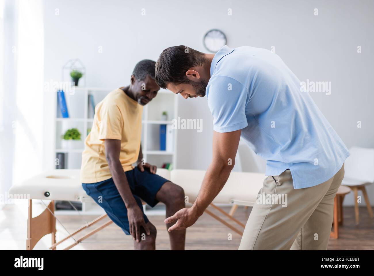 Junger Rehabilitologe, der mit der Hand auf das Knie eines afroamerikanischen Patienten zeigt Stockfoto