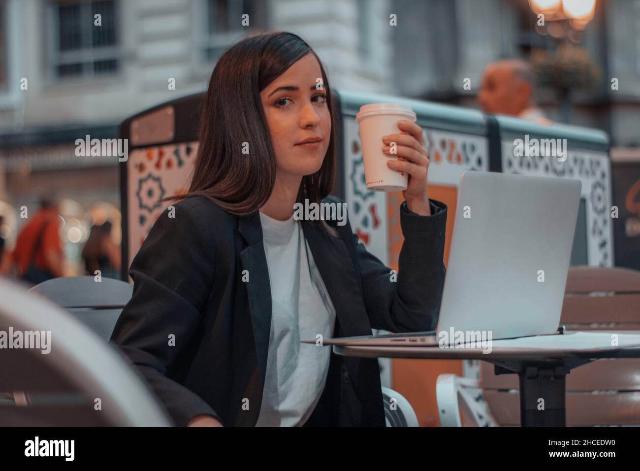 Die faszinierte Lehrerin, die am frühen Morgen bei Sonnenaufgang in einer Cafeteria mit ihrem Laptop-Computer Kaffee trinkt und auf einem Stadtplatz ein glückliches Lächeln auf den Lippen hat Stockfoto