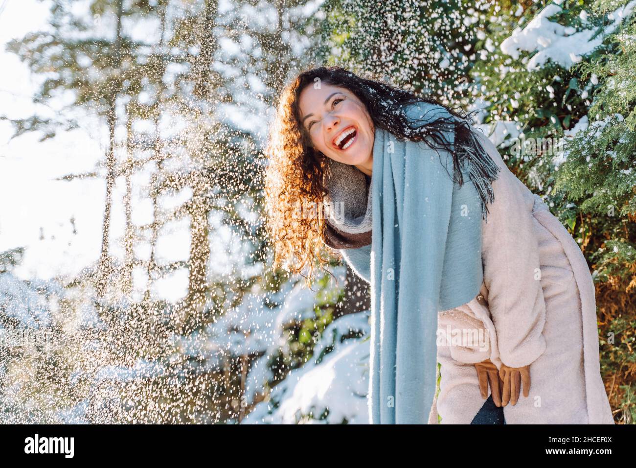 Lächelnd attraktive junge Frau mit langen Haaren haben Spaß unter fallendem Schnee tragen warme Kleidung und blauen Schal in verschneiten Winterwald. Genießen Sie die Natur, Stockfoto