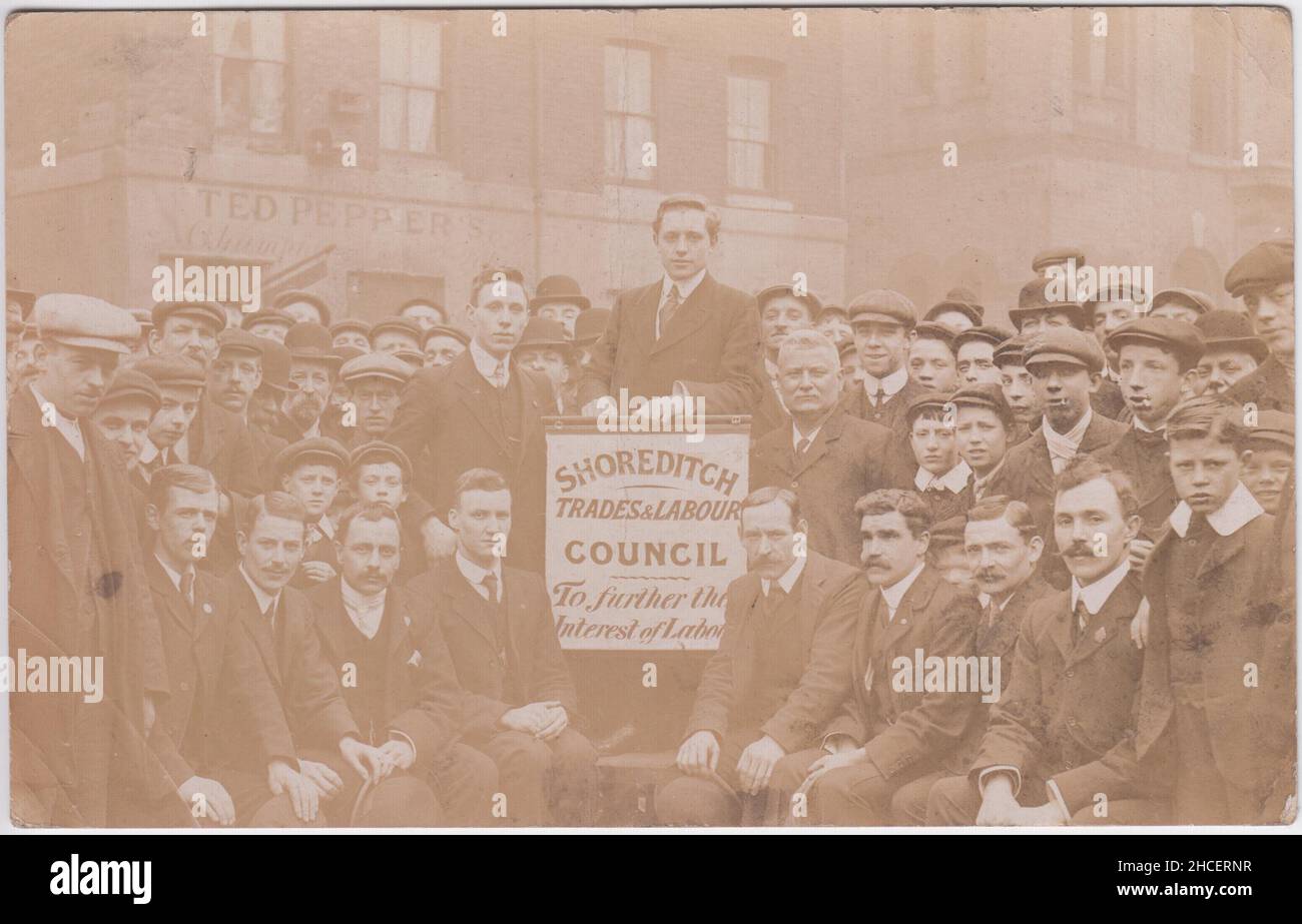 Shoreditch Trades und Labour Council. Um das Interesse der Arbeit zu fördern', ca. 1910: Gruppe von Männern auf den Straßen von Shoreditch, London, außerhalb der Räumlichkeiten von Ted Pepper's. Ein Mann hält ein kleines Banner des Shoreditch Trades & Labour Council. Viele der Zuschauer sind jung und nicht wenige tragen flache Kappen. Das Bild befindet sich auf einer Fotopostkarte, die im Januar 1910 gesendet wurde, und kann sich damit auf den allgemeinen Wahlkampf beziehen Stockfoto