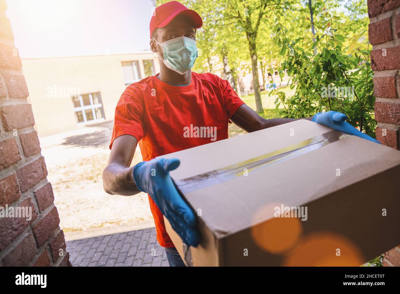 Delivery Mann Mitarbeiter in roten Kappe blank T-Shirt Uniform Gesichtsmaske Handschuhe halten leere Karton-Box. Service Quarantäne Pandemie Coronavirus Virus 2019 Stockfoto