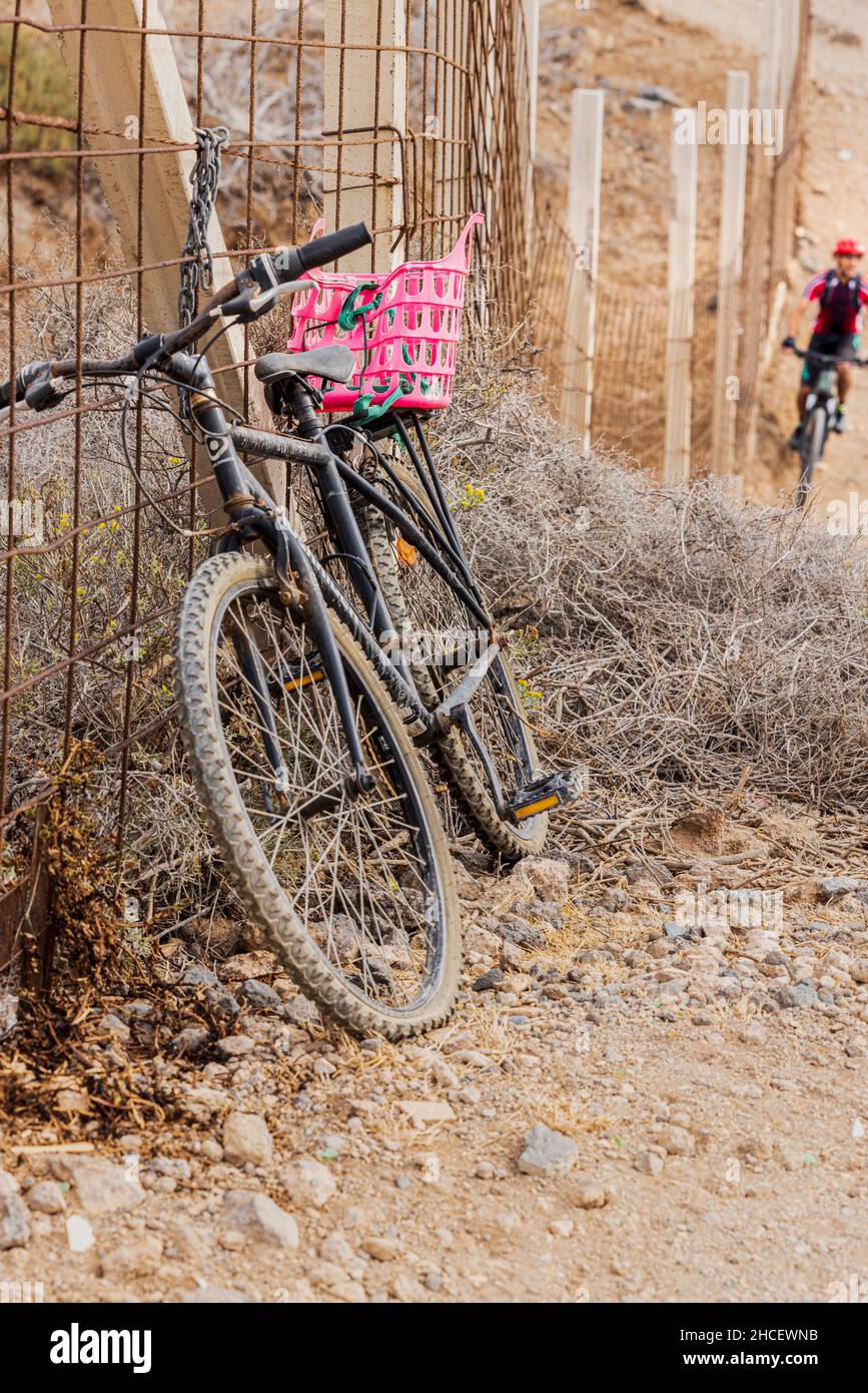 Altes Fahrrad, das an einen Drahtzaun mit einem roten Plastikkorb auf der Rückseite gekettet ist, als sich ein Mountainbiker von hinten nähert, in der Nähe von Los Abrigos, Teneriffa, Stockfoto
