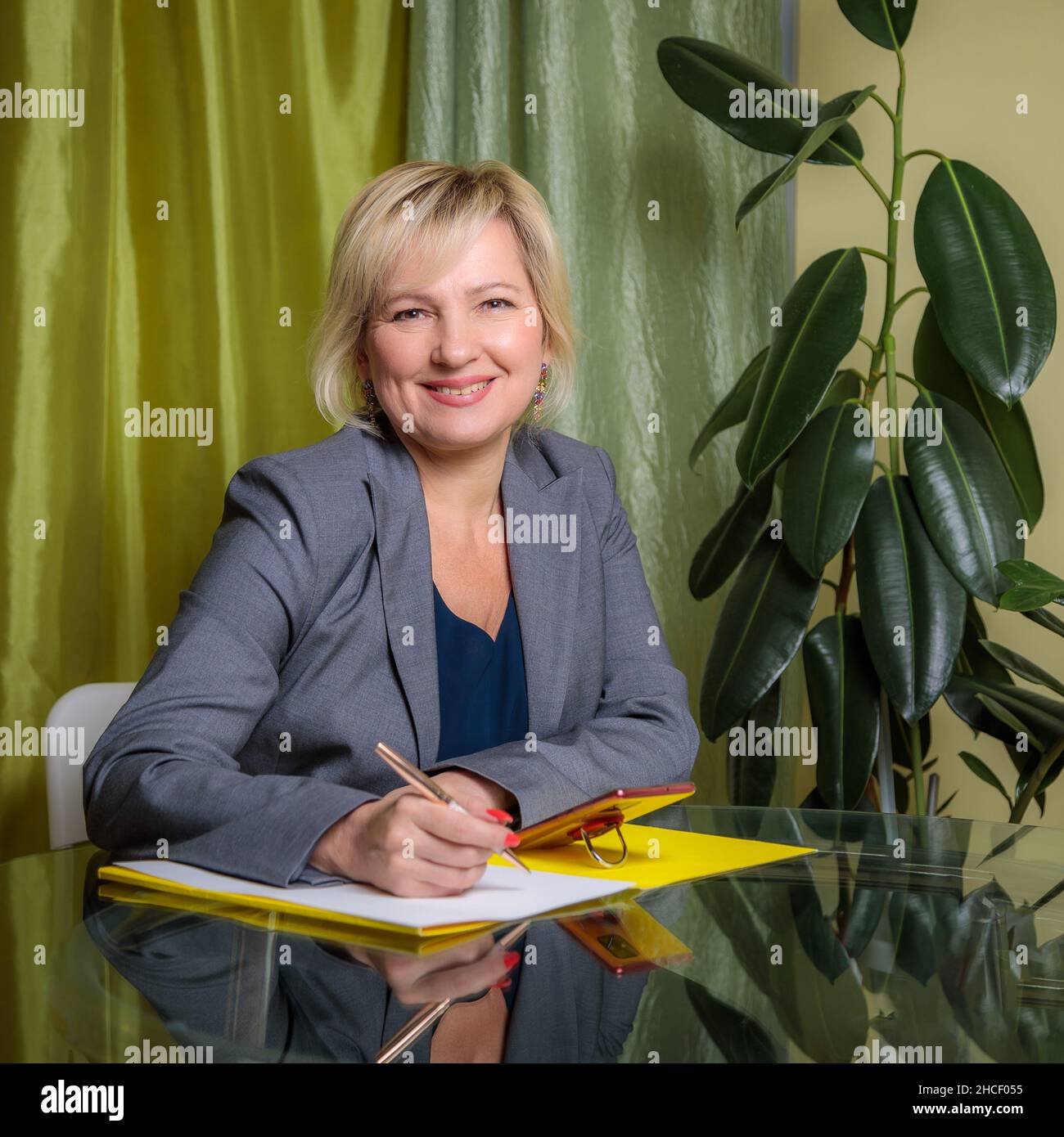 Eine attraktive Frau mittleren Alters, die Regisseurin, sitzt an einem Tisch im Büro und füllt Dokumente aus. Lächelt für die Kamera. Stockfoto