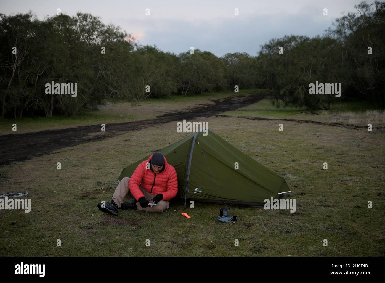 Wilde Lager- und Überlebenskünste bei Outdoor-Aktivitäten schärft ein Mann einen hölzernen Ast, der vor dem Zelt sitzt Stockfoto