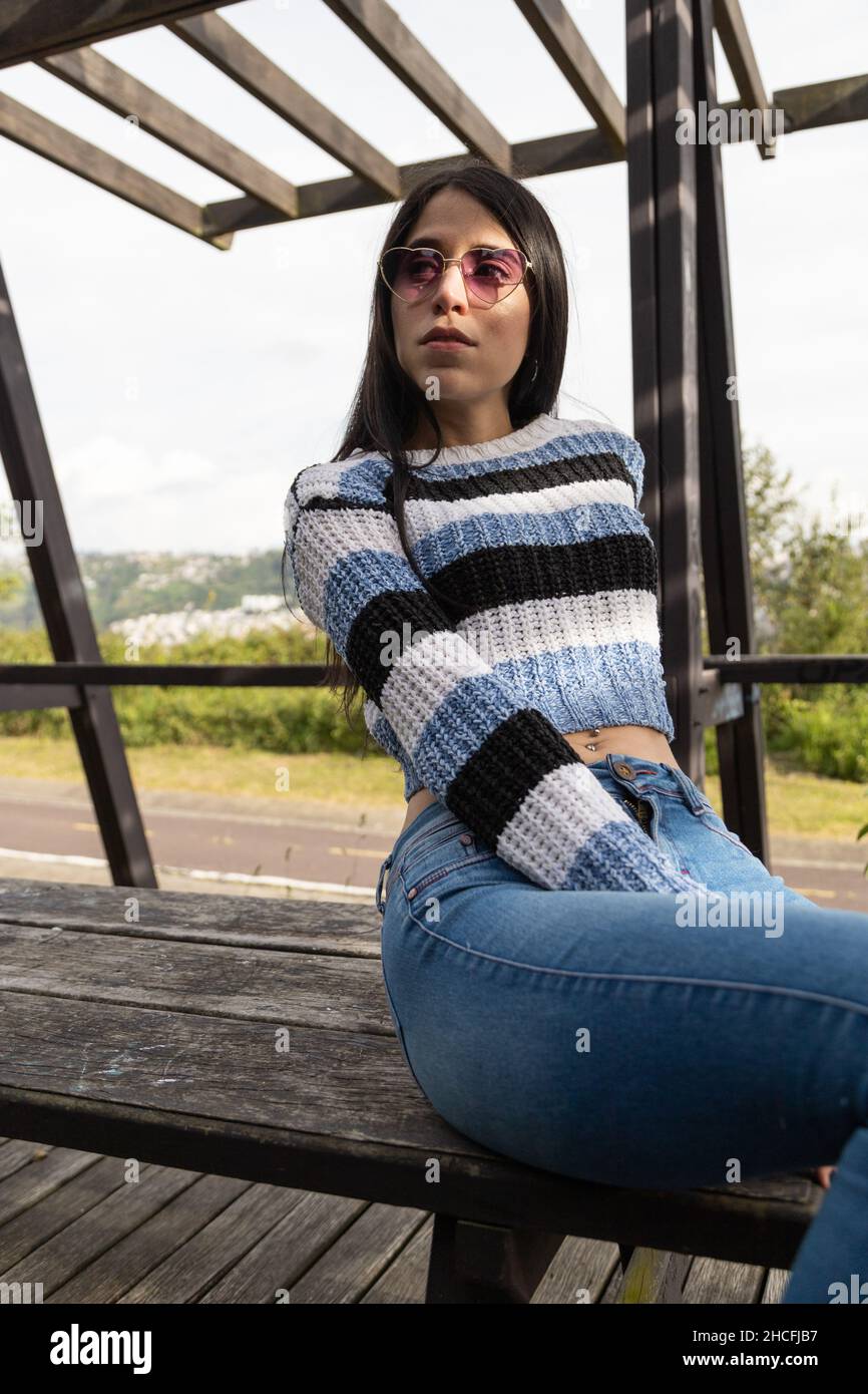 Schönes junges lateinisches Model, das mit schwarzen, geraden Haaren in einem Park mit Natur posiert, trägt Sonnenbrille, Jeans und lässigen Pullover, Schönheit und Mode Stockfoto