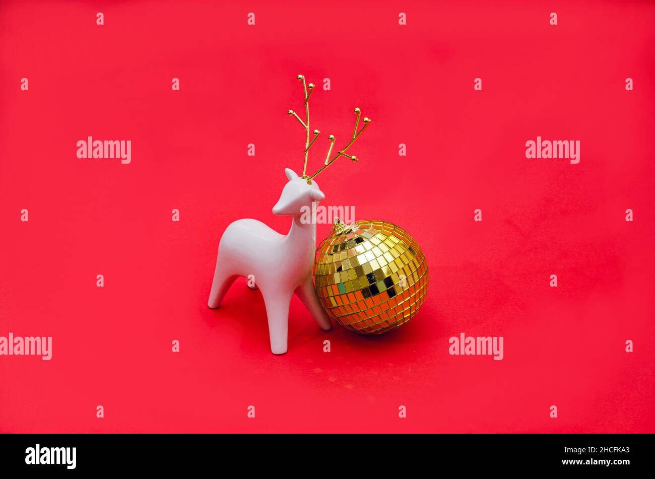 Ein weißes Rentier mit goldenen Hörnern neben einem großen goldenen Discokugel-Ornament auf hellrotem Hintergrund. Kreatives Konzept für festliche Winterpartys. Surr Stockfoto