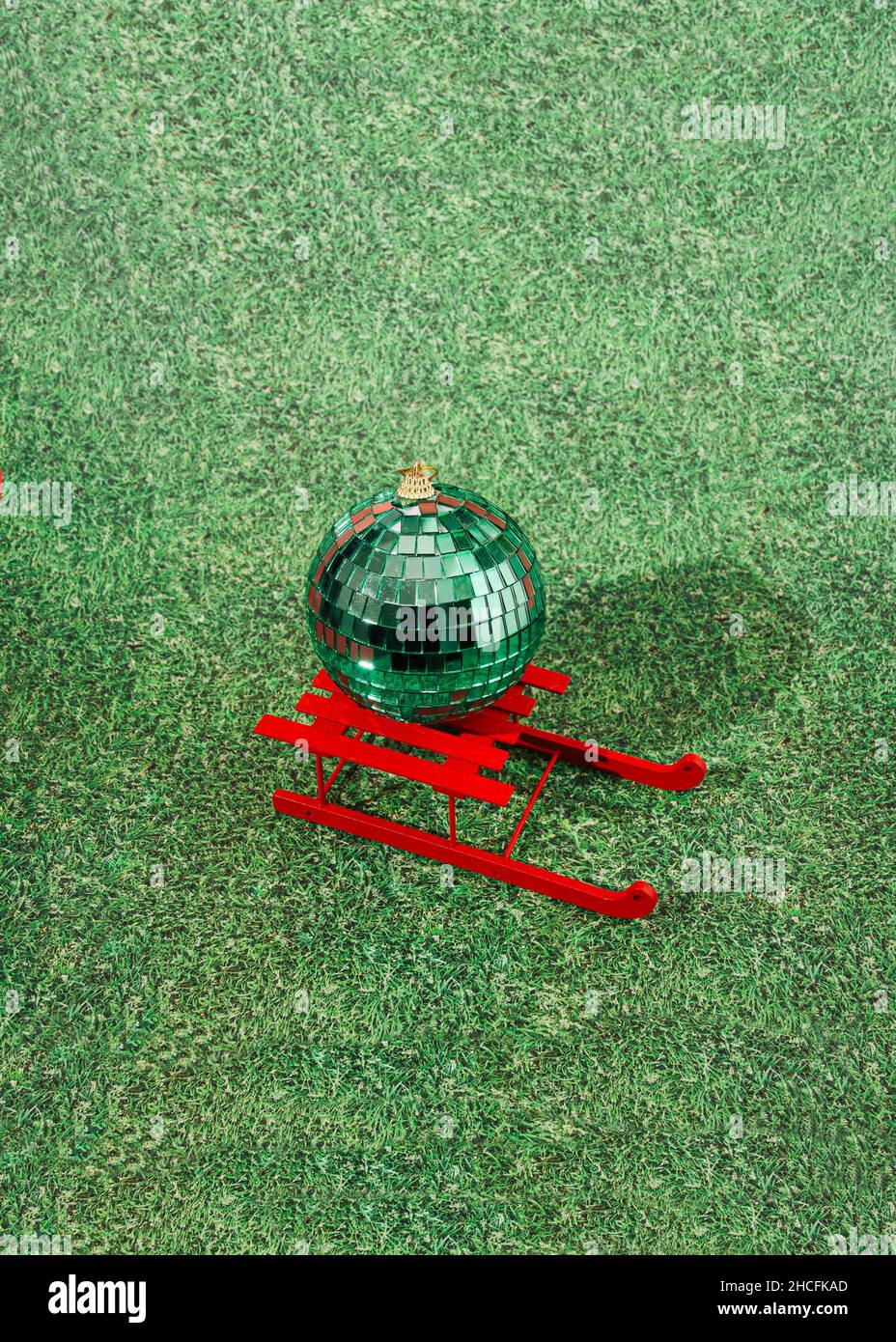 Eine riesige grüne Disco Ball Ornament auf roten Schlitten auf Kunstrasen Hintergrund. Kreatives Konzept für festliche Feiertage an einem warmen Ort. Design für Adve Stockfoto