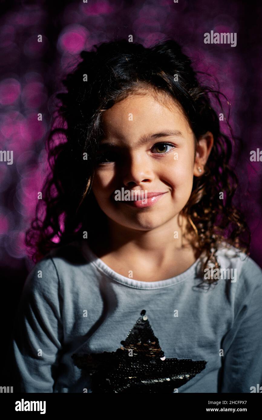 Bezauberndes lockiges Spanisches Mädchen lächelt, während es vor einem verschwommenen violetten Hintergrund auf die Kamera blickt Stockfoto