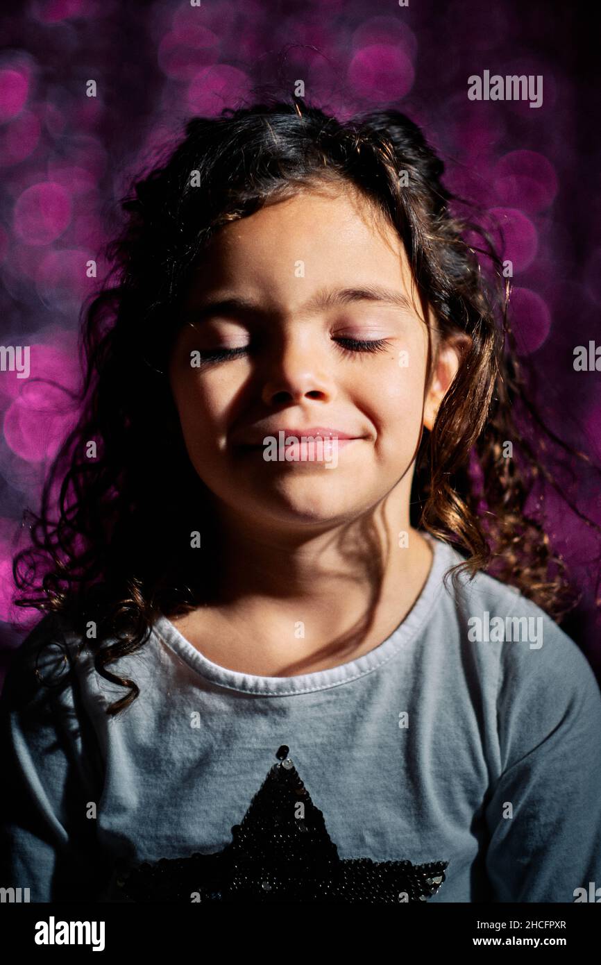 Liebenswert lockiges spanisches Mädchen lächelt, während sie ihre Augen vor einem verschwommenen violetten Hintergrund schließt Stockfoto