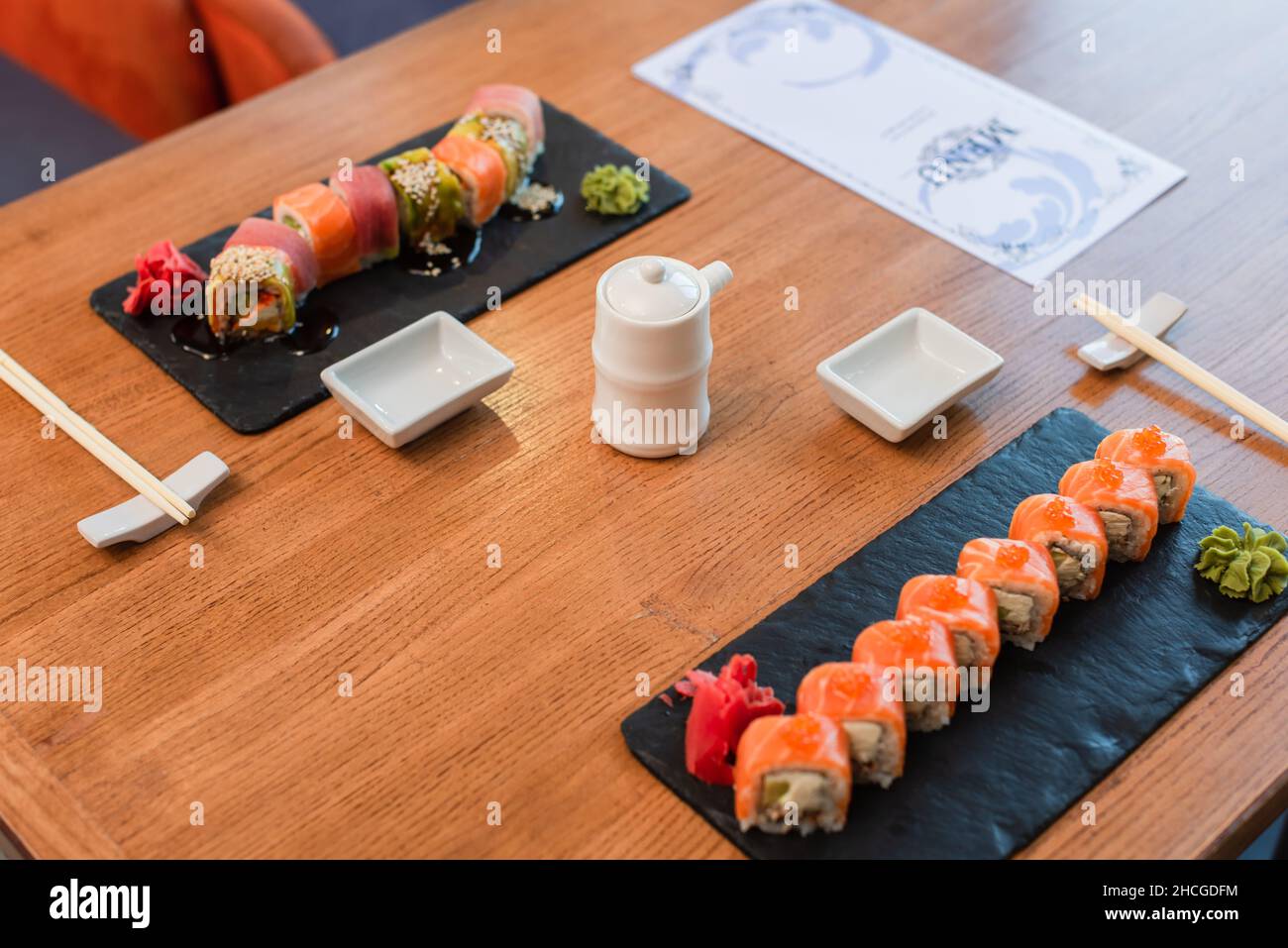 Schwarze Teller mit leckeren Sushi-Rollen mit Wasabi und Ingwer in der Nähe von Sojasauce-Topf und Menübroschüre auf Holztisch Stockfoto