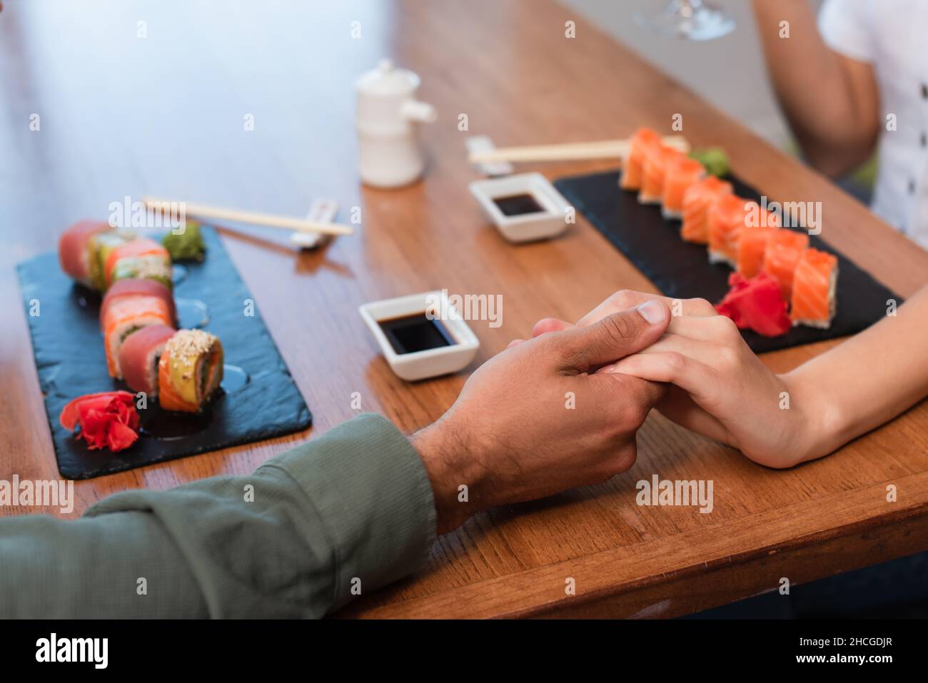Beschnittene Ansicht eines Paares, das seine Hände in der Nähe von Sushi-Brötchen und Schüsseln mit Sojasauce auf dem Tisch hält Stockfoto