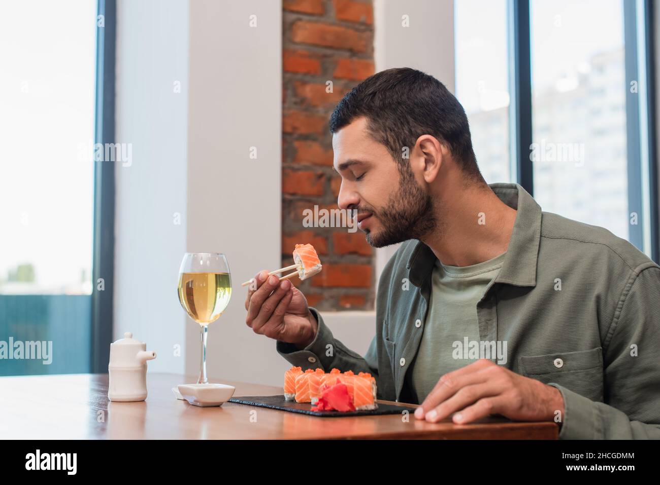 Junger Mann mit geschlossenen Augen hält köstliches Sushi mit Essstäbchen in der Nähe von Glas oder Weißwein Stockfoto