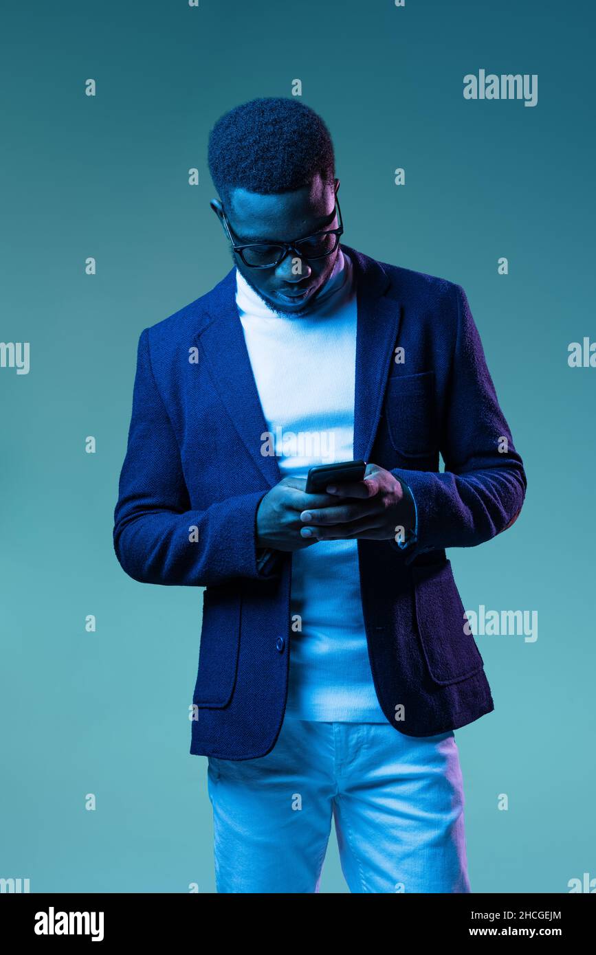 Halblanges Porträt eines jungen, stylischen afrikanischen Mannes, Studenten mit weißem Nackenpolo und Jacke auf blauem Hintergrund in Neonlicht isoliert. Stockfoto
