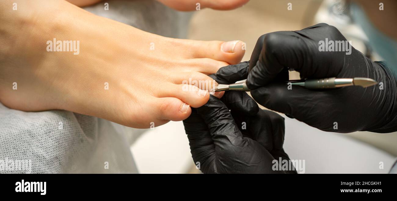 Nagelhautentfernung an den Zehen. Hände in schwarzen Handschuhen des Pediküre-Meisters entfernen die Nagelhaut an den weiblichen Zehen durch Stopfer Stockfoto