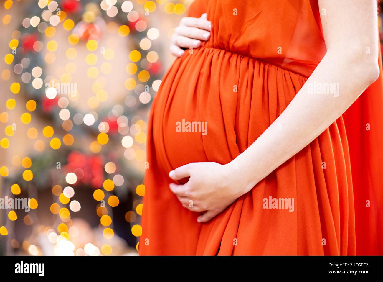 Bestes weihnachtsgeschenk. Schwangere in einem roten Kleid umarmt ihren Bauch vor dem Hintergrund des Weihnachtswohnzimmers in Unschärfe. Nahaufnahme kein Gesicht. Stockfoto