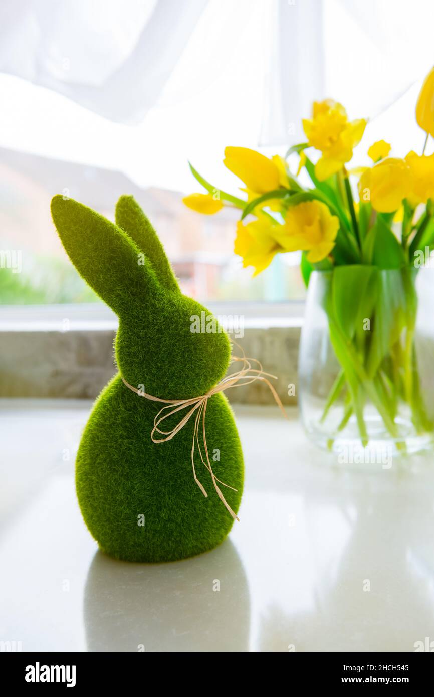 Niedliche grüne Gras Hase Kaninchen und Bouquet von frischen Frühling gelben Tulpen und Narzissen Blumen auf dem weißen Küchentisch in der Nähe des Fensters. Frohe Ostern Stockfoto