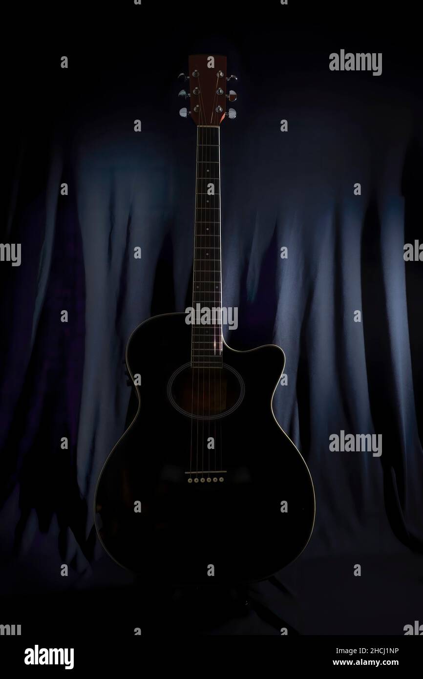 Low-Key-Bild der Silhouette einer akustischen Gitarre auf einer Bühne mit einem leicht beleuchteten schwarzen Hintergrund Vorhang im Hintergrund Stockfoto