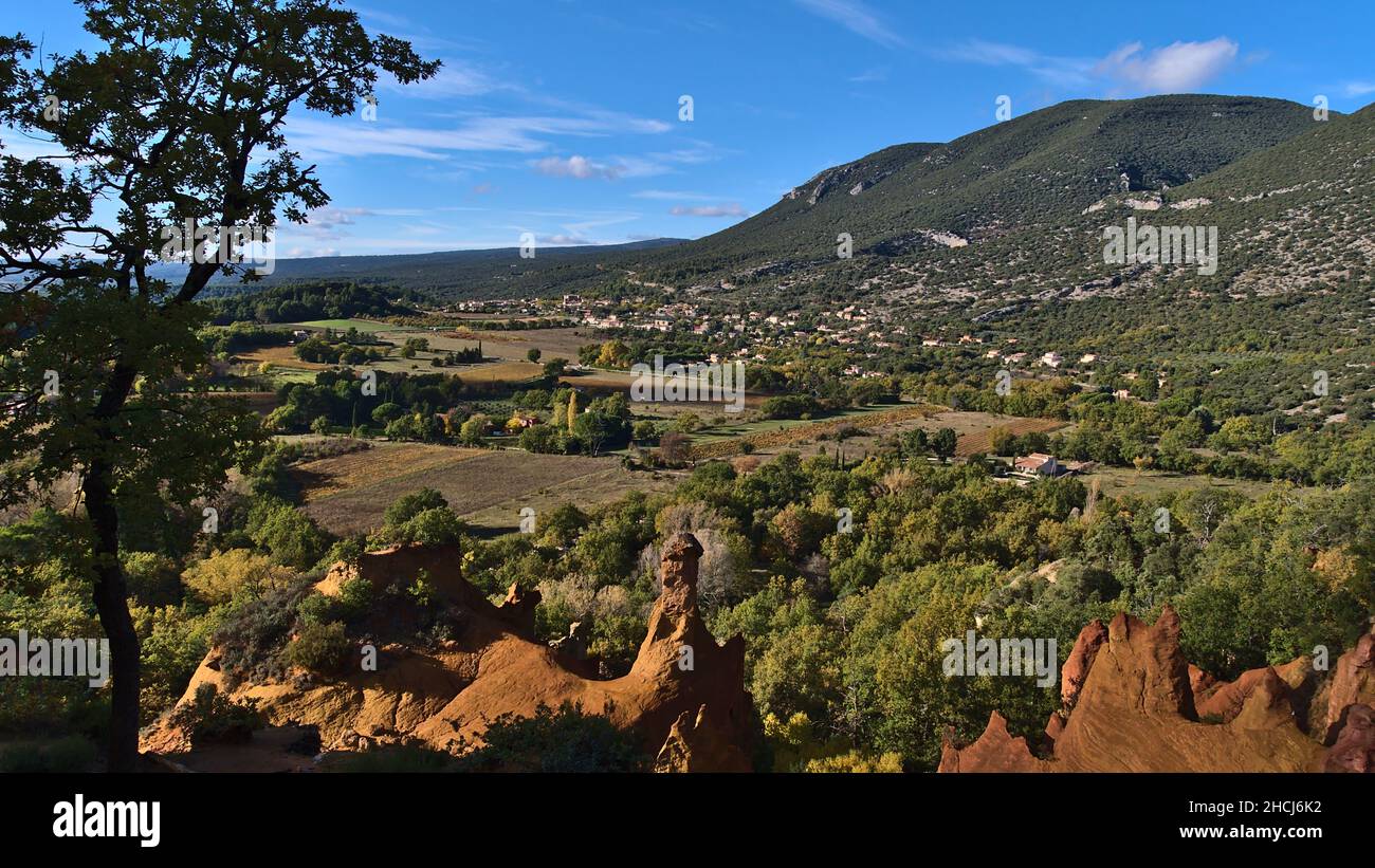 Schöne Aussicht auf das Luberon-Tal mit bunten ockerfarbenen Felsen bei Colorado Provenzal mit Rustrel-Dorf im Hintergrund in der Provence, Frankreich. Stockfoto