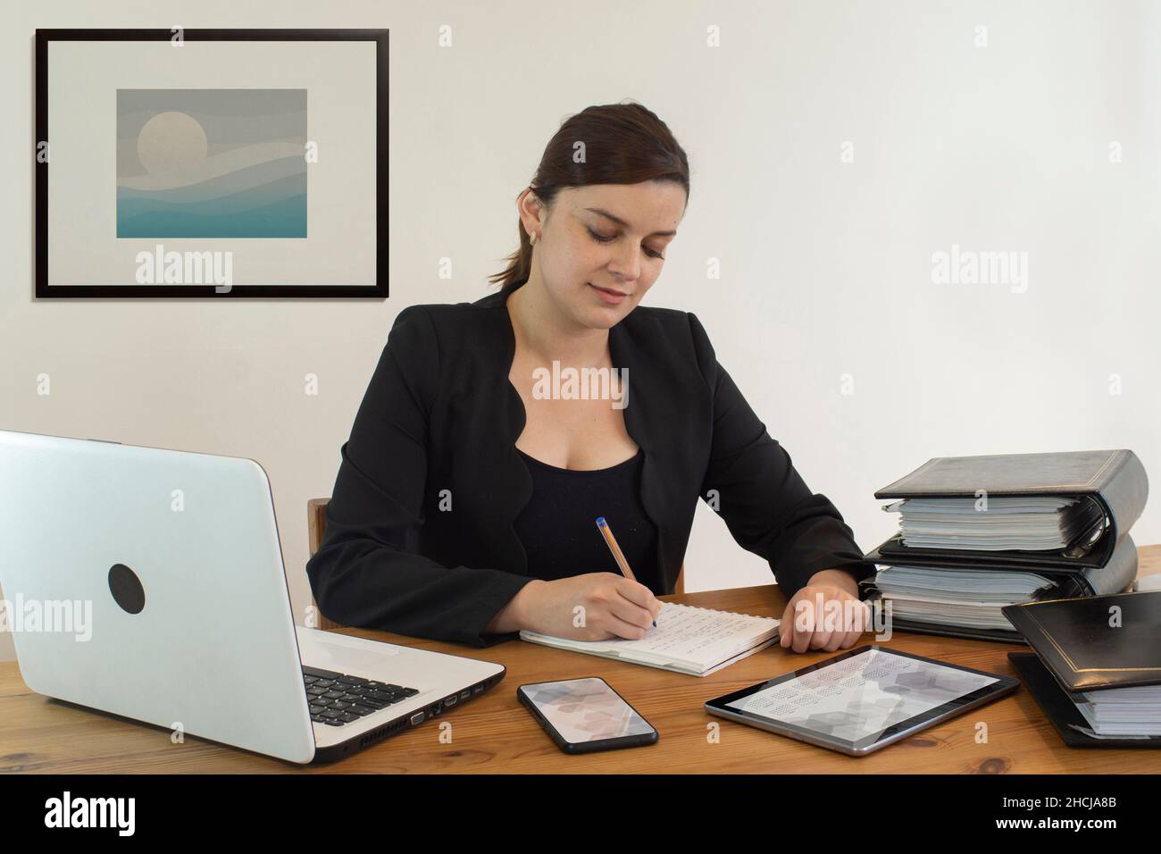 Schöne hispanische Angestellte, die glücklich lächelte und an ihrem Schreibtisch saß und mit dem Schreiben in ihrem Notebook gegen die weiße Wand beschäftigt war Stockfoto