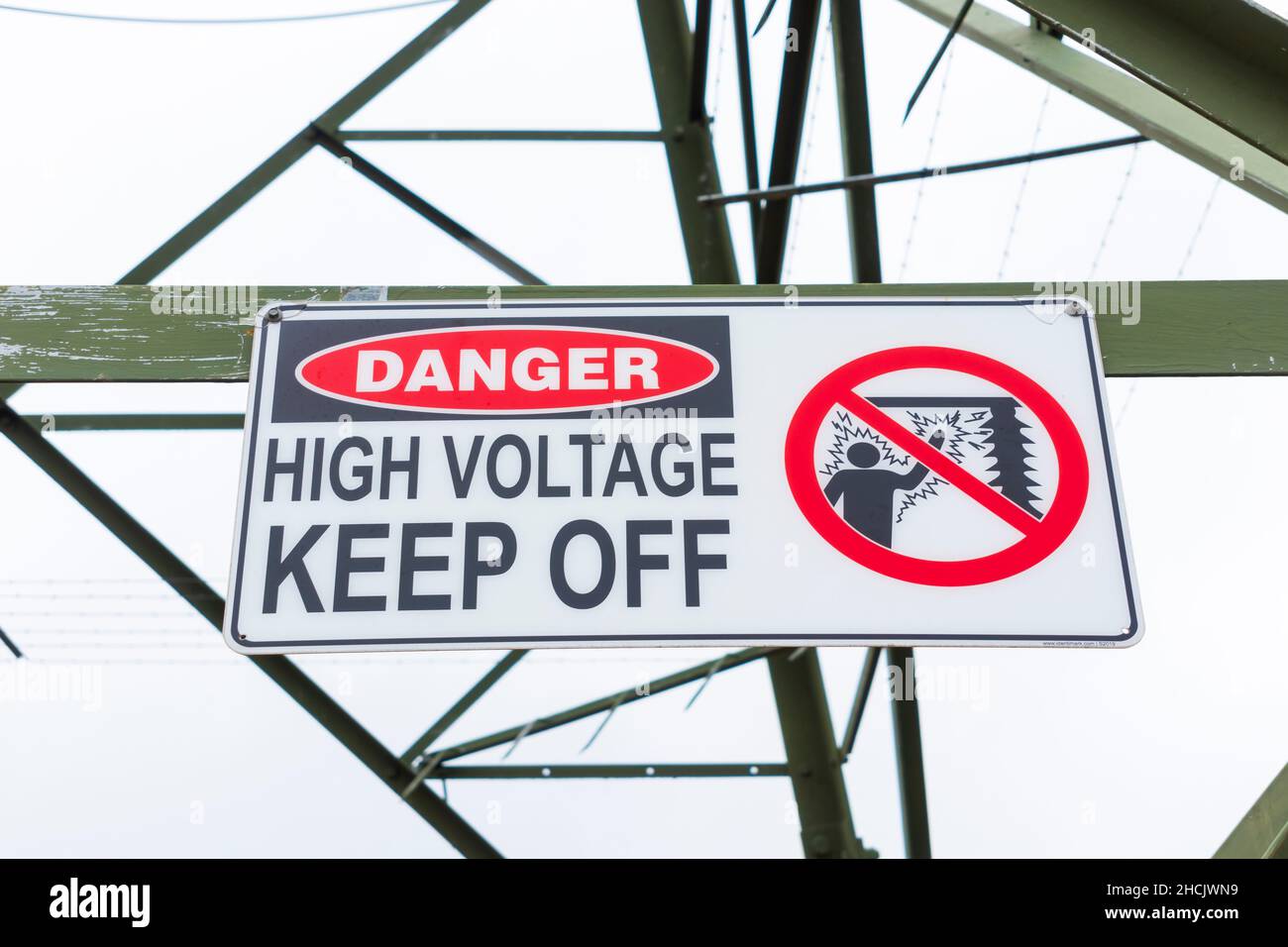 Foto eines Sicherheitszeichens mit Hochspannungsfreischaltung auf einem großen elektrischen Übertragungsturm aus Stahl vor einem grau bewölkten Himmel Stockfoto