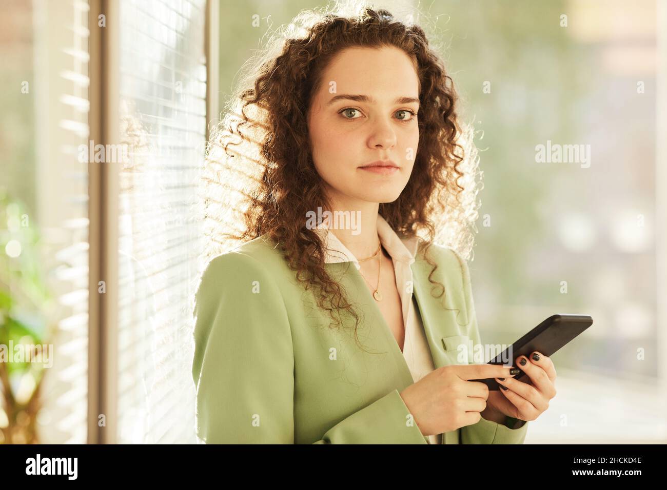 Horizontale, mittlere Nahaufnahme einer attraktiven jungen kaukasischen Frau, die im modernen Büro mit dem Smartphone und der Kamera steht Stockfoto