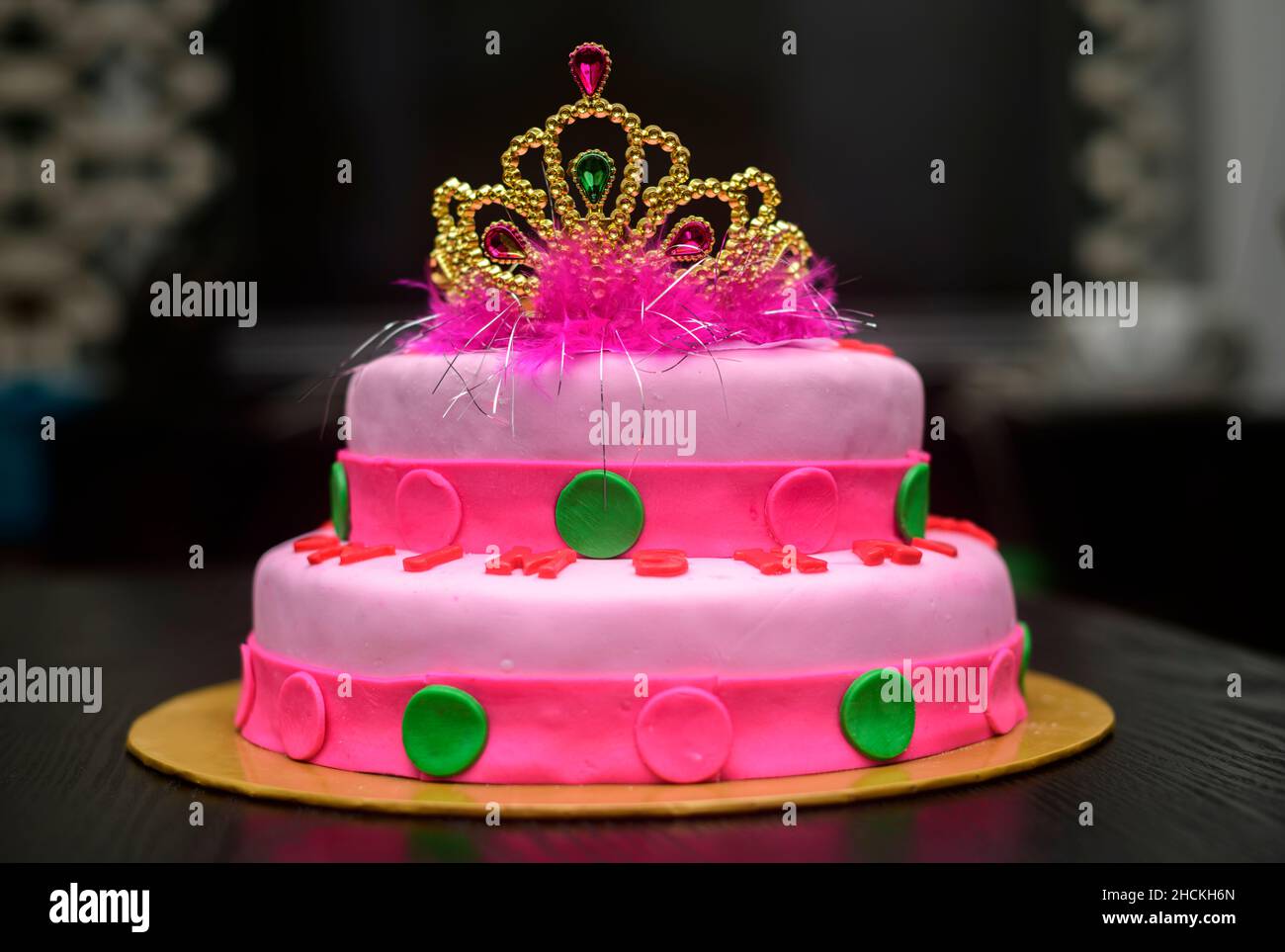 Zwei-Tier-Fondant-Kuchen-Design mit einer Tiara auf der Oberseite, mit Puderblumen verziert. Köstliche rosa-rote farblich thematisierte runde Torte auf gelbem Kuchenblech dagegen Stockfoto