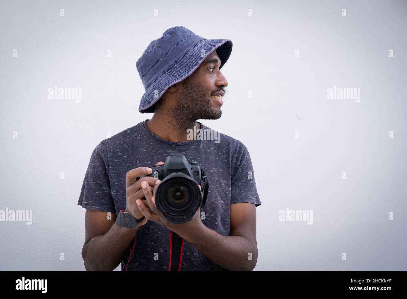 Ein professioneller Fotograf hält eine Kamera in der Hand. Ein stilvoller und attraktiver Mann trägt einen Eimer Hut und lächelt, während er eine DSLR hält, er ist isoliert vom Whi Stockfoto