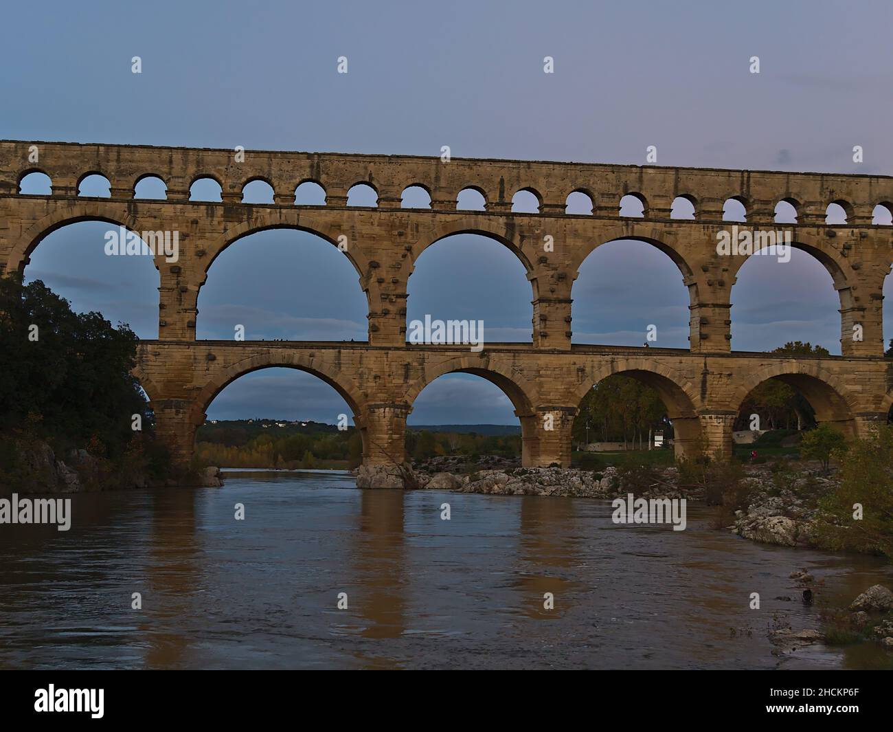 Blick auf das antike römische Aquädukt Pont du Gard, das am Abend den Fluss Gardon überspannt, in der Nähe der Stadt Vers-Pont-du-Gard, Ockitanie, Frankreich mit Felsen. Stockfoto