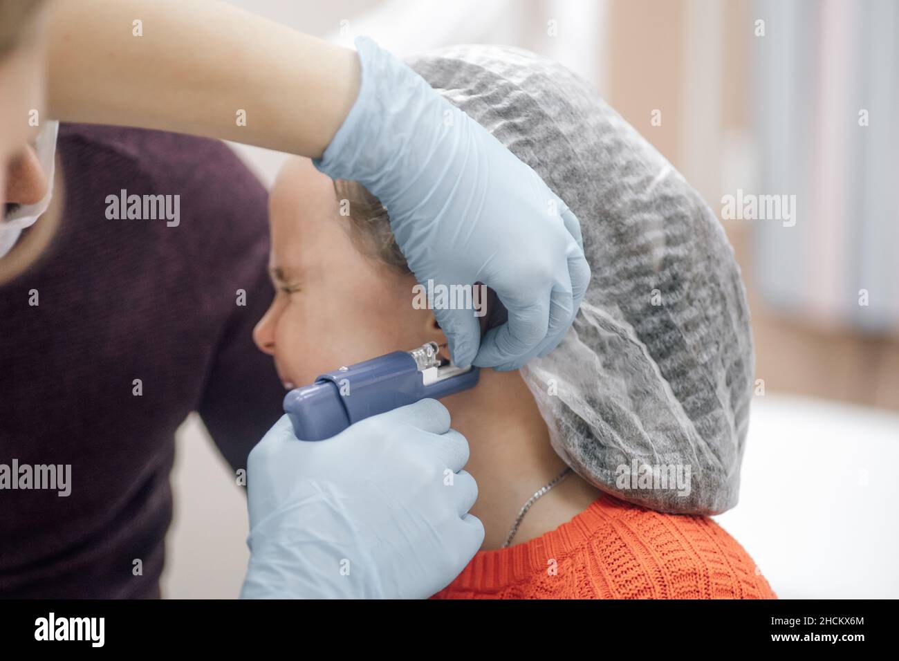 Nahaufnahme der Krankenschwesternhände in medizinischen Einweghandschuhen durchdringt Ohr mit Pistole für stechende Ohren des Kindes. Kleines Mädchen in Einweg-medizinische Kappe weint in Stockfoto