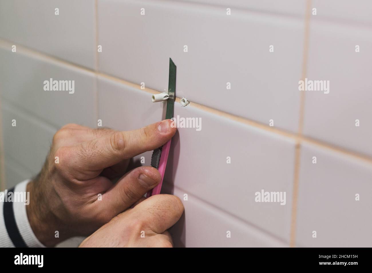 Nahaufnahme von Männerhänden, die im Waschraum mit einem Schreibwarenmesser einen zusätzlichen Teil der Dübel in die geflieste Wand einschneiden. Im Bad wurden Renovierungsarbeiten durchgeführt. Schmutz von Stockfoto