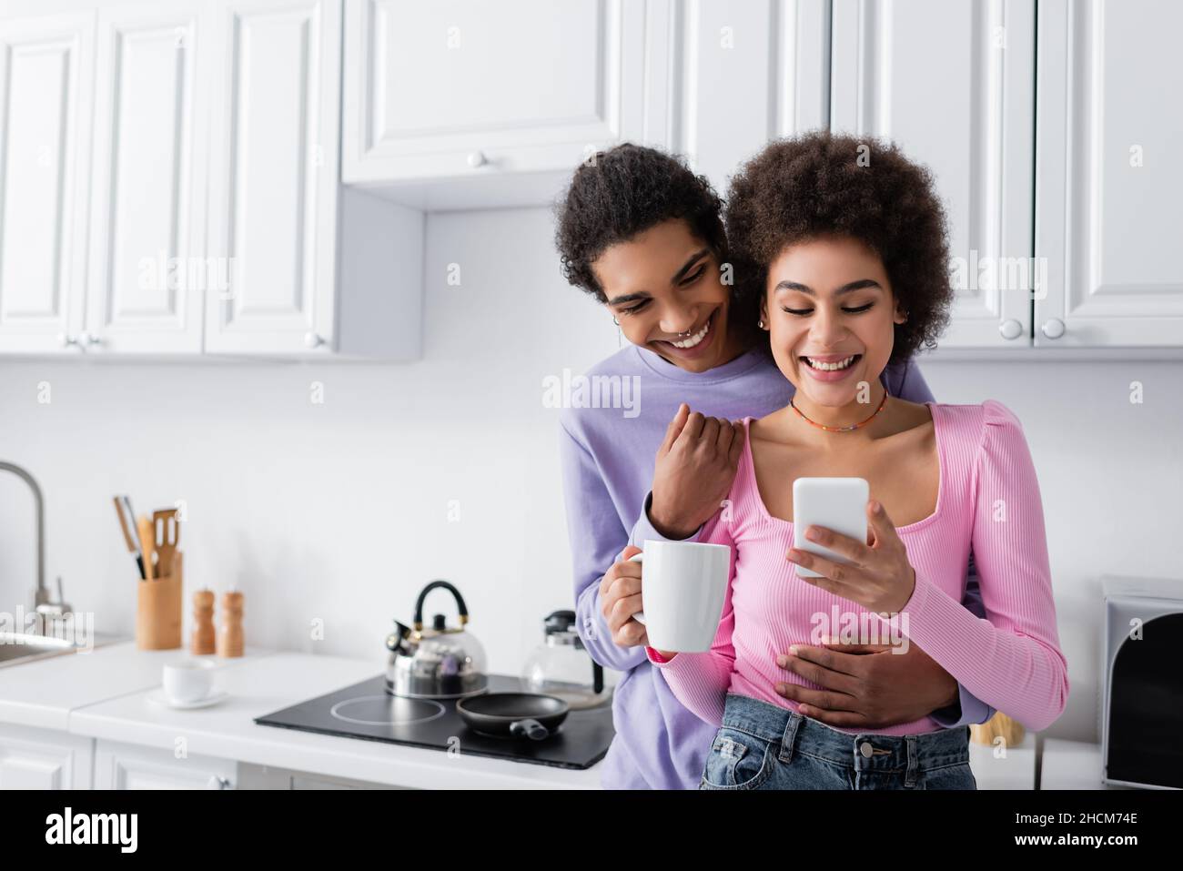 Positive afroamerikanische Frau mit Tasse mit Smartphone in der Nähe von Freund zu Hause Stockfoto