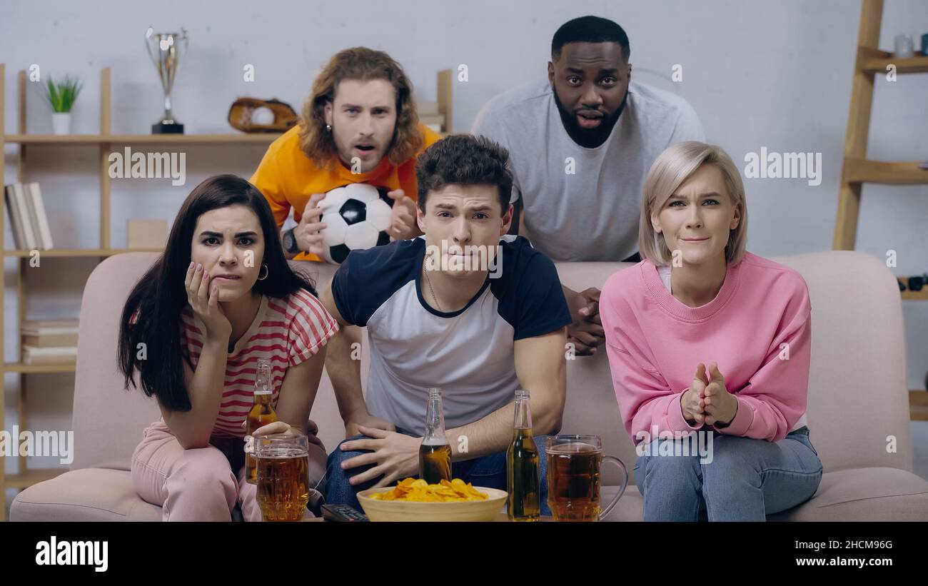 Ängstliche multikulturelle Sportfans, die sich ein Fußballspiel in der Nähe von Bier und Chips zu Hause ansehen Stockfoto