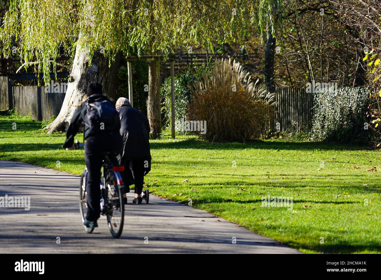 Eine ältere Dame in einem schwarzen Mantel geht auf einem Bürgersteig in einem Park und lehnt sich auf ihren Spaziergänger. Ein Radfahrer fährt an ihr vorbei. Stockfoto