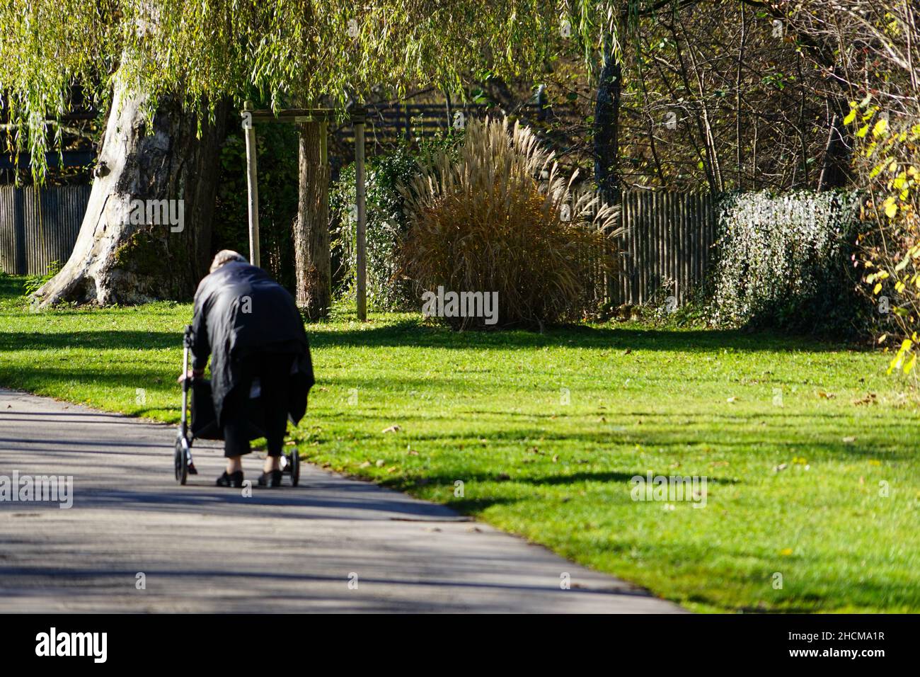 Eine ältere Dame in einem schwarzen Mantel geht auf einem Fußweg in einem Park und lehnt sich an ihren Spaziergänger. Stockfoto
