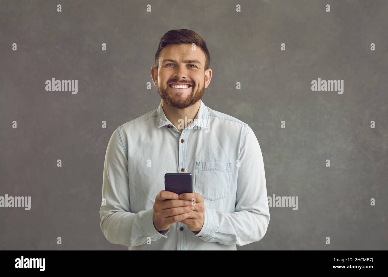 Glücklicher junger Mann mit Smartphone und lächelnd, isoliert auf grauem Hintergrund stehend Stockfoto