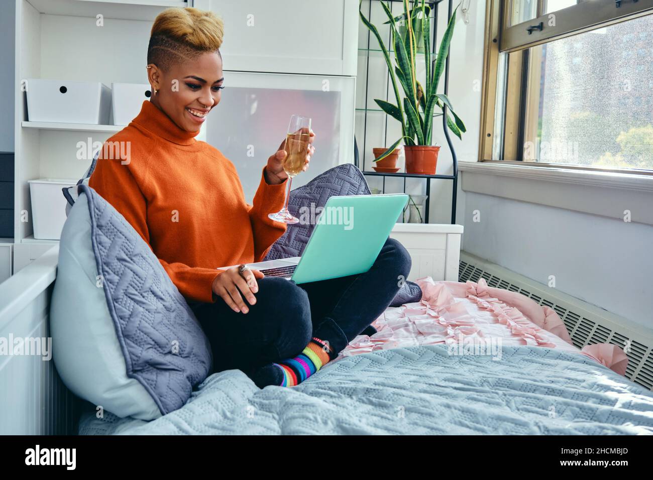 Lächelnde junge Hipsterin, die Champagnerflöte zeigt, während sie zu Hause über einen Laptop per Video anruft Stockfoto