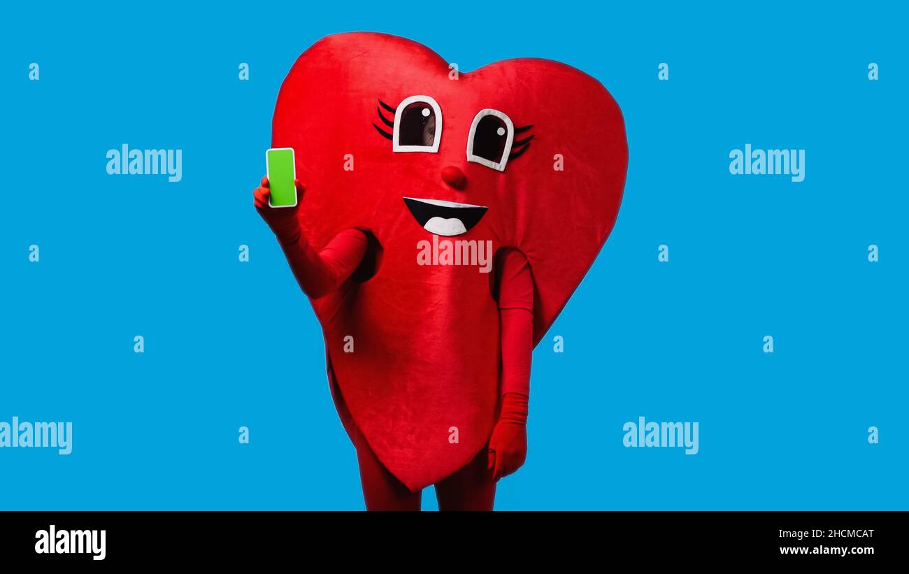 Person im positiven Herzkostüm hält Smartphone mit grünem Bildschirm isoliert auf blau Stockfoto