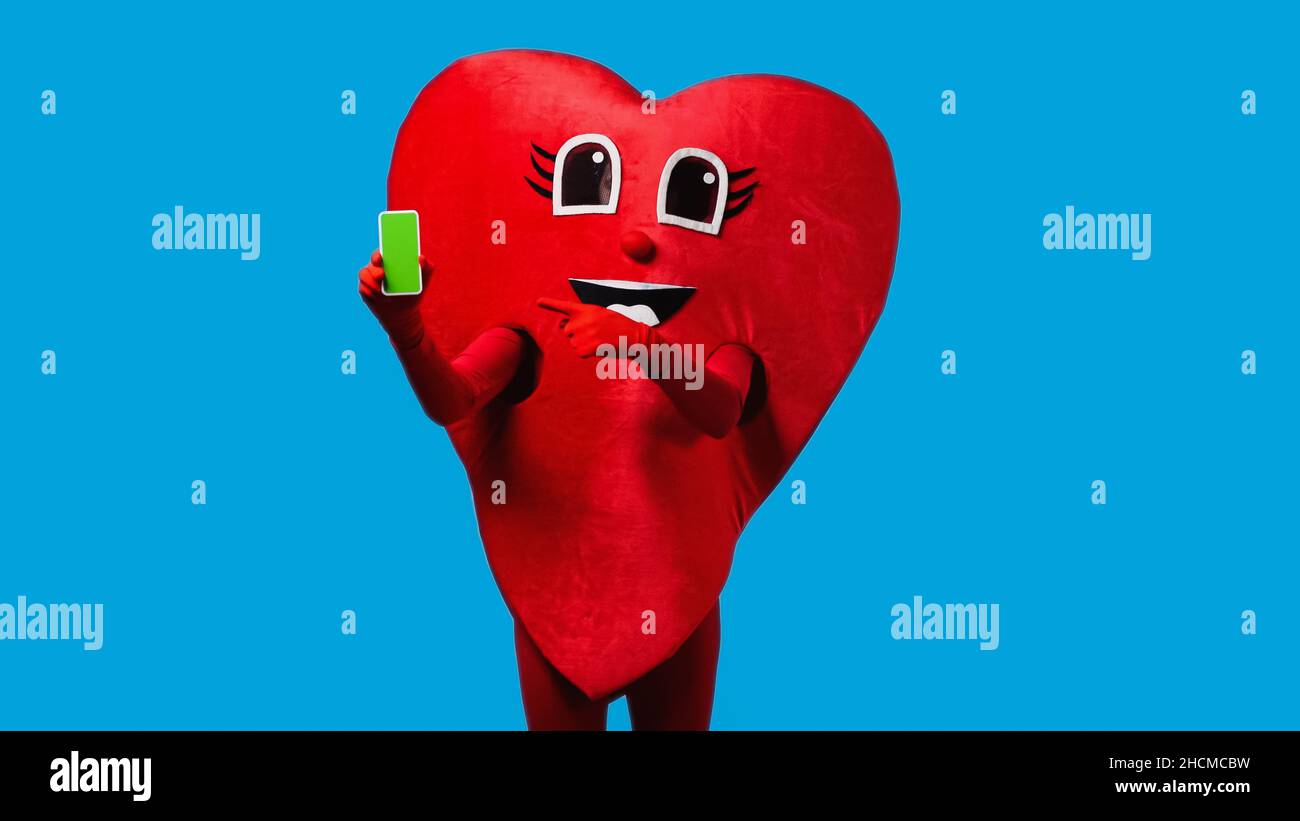 Person im positiven Herzkostüm, die auf das Smartphone zeigt, mit einem grünen Bildschirm, der auf Blau isoliert ist Stockfoto