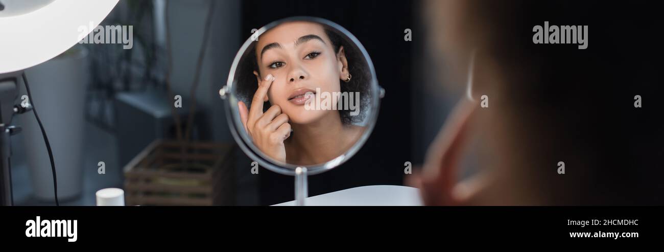 Junge afroamerikanische Frau, die kosmetische Creme in der Nähe des Spiegels und Ringlicht zu Hause, Banner Stockfoto