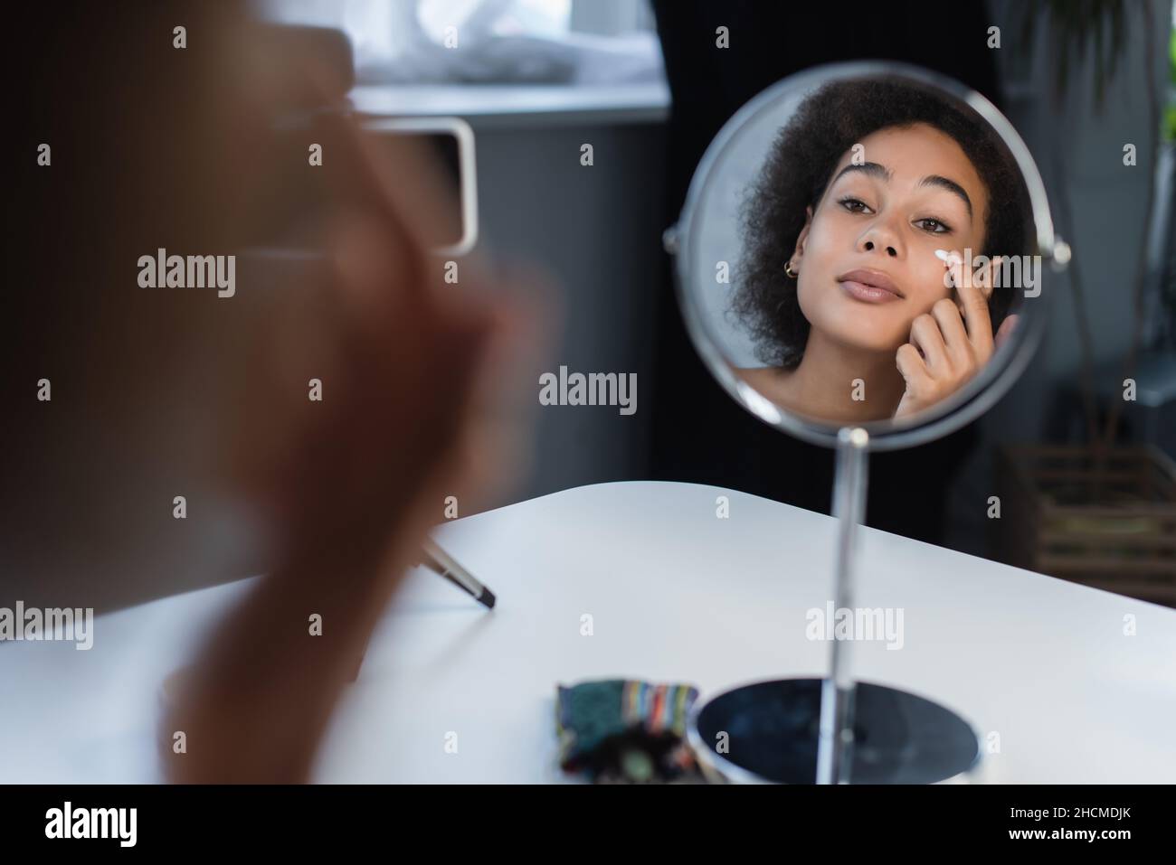 afroamerikanische Bloggerin, die zu Hause Kosmetikcreme auf das Gesicht in der Nähe von Spiegel und Smartphone anwendet Stockfoto