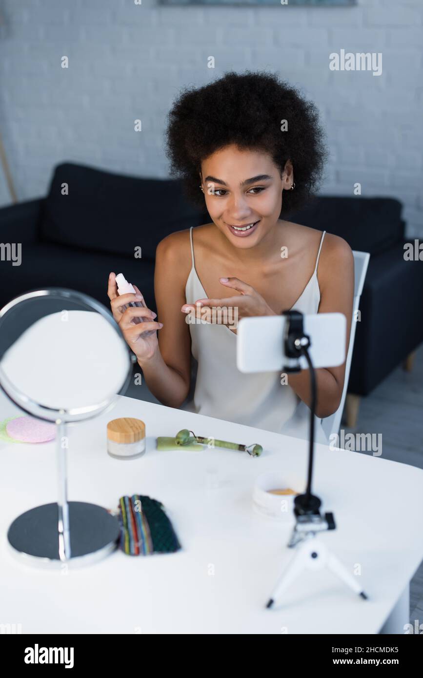 Lächelnder afroamerikanischer Blogger zeigt auf Kosmetiklotion in der Nähe von Smartphone und Spiegel zu Hause Stockfoto