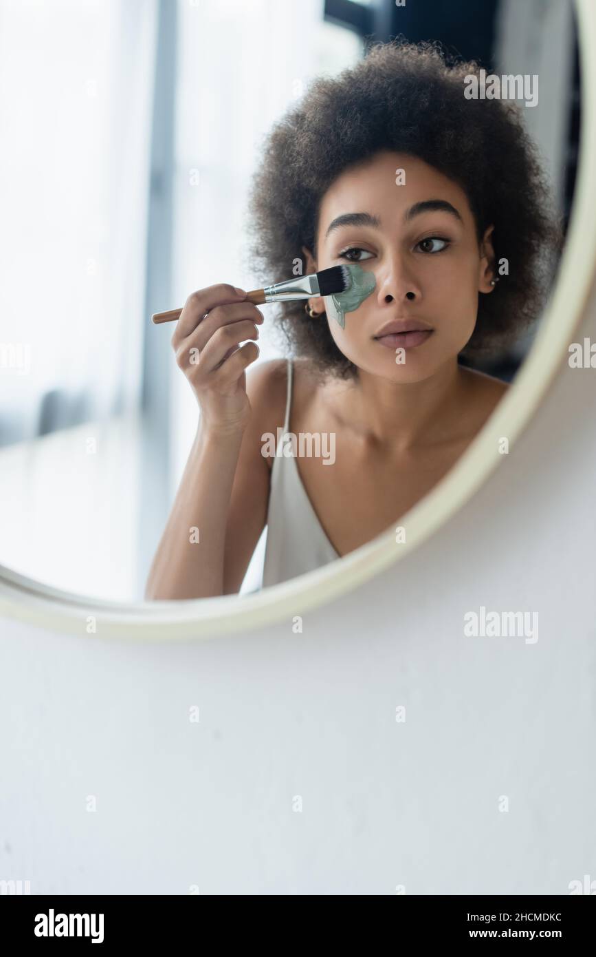 afroamerikanische Frau, die Gesichtsmaske mit Kosmetikpinsel in der Nähe des Spiegels zu Hause anwendet Stockfoto