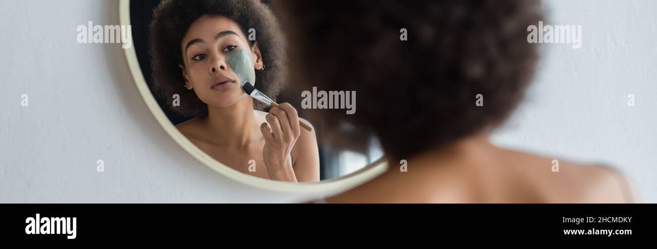 Junge afroamerikanische Frau mit kosmetischen Pinsel Anwendung Gesichtsmaske in der Nähe von Spiegel, Banner Stockfoto