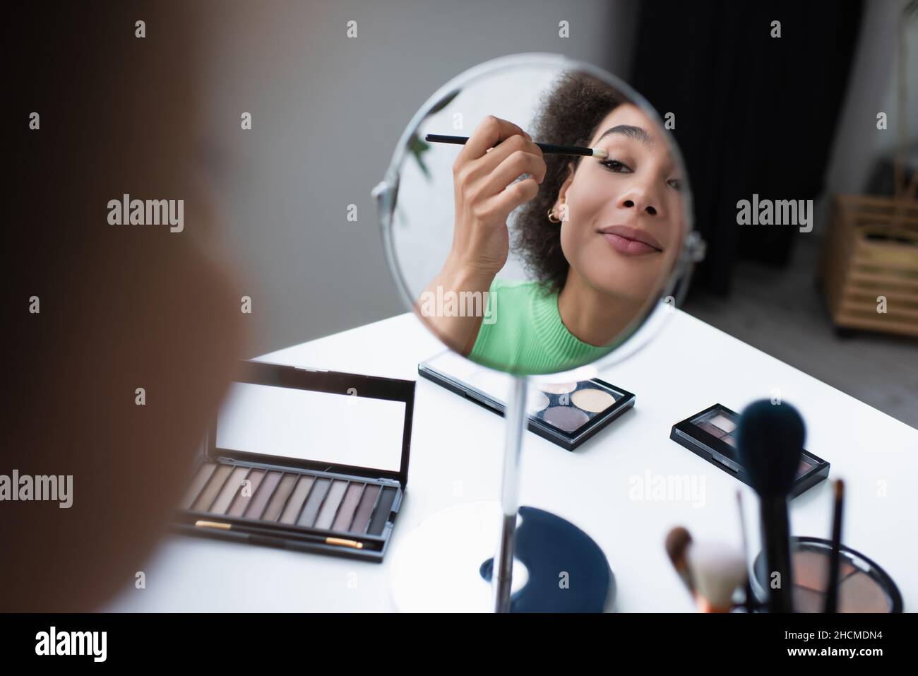 Lächelnde afroamerikanische Frau, die Augenschatten in der Nähe des Spiegels und verschwommene Kosmetikbürsten zu Hause anwendet Stockfoto